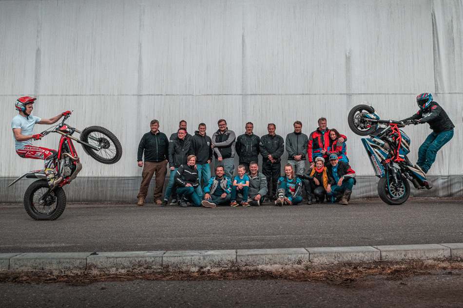 Tým a účastníci motoškoly u betonové zdi, po stranách jezdci na trialce a supermotu na zadním kole